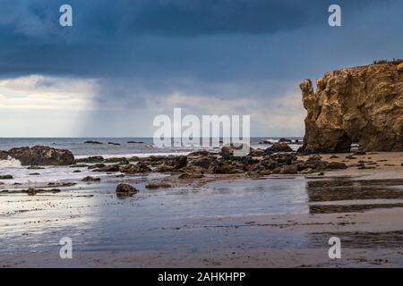 Coastline view of tall bluffs along El Matador Beach with reflections in the beach water and dramatic rain clouds in the distance, Malibu, California Stock Photo