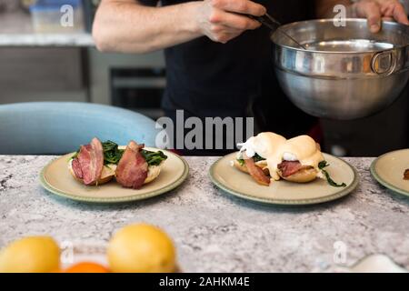 Eggs benedict with crisp bacon and spinach being topped with hollandaise sauce for morning brunch. Stock Photo