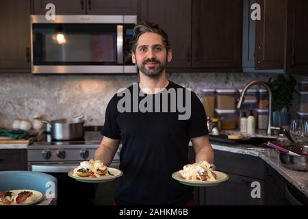 A ethnically mixed male homecook in a kitchen holding plates of eggs benedict. Stock Photo