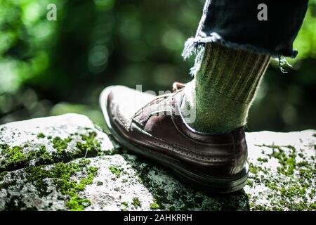 Stepping on the edge of a rock covered with green moss. Confident young male wearing good looking shoes with green hippie wool socks and blue torn jeans. Stock Photo