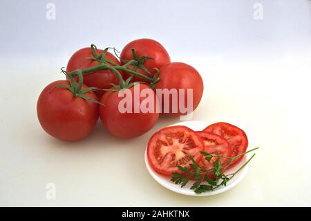 A plate with sliced tomatoes and a sprig of fresh parsley next to a few tomatoes on a branch on a white neutral background Stock Photo