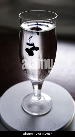 Blue and black ink dropping into a wine glass of water on a white background. Stock Photo