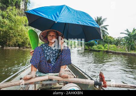 An elderly woman uses her feet to push the oars of a rowboat, while holding a fan and umbrella, taking tourists down the Ngo Dong River, Vietnam Stock Photo