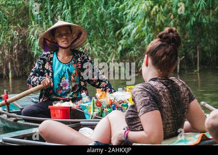 A woman tries to sell candy from her boat to a tourist on another boat in the Ngo Dong River, Ninh Binh Province, Vietnam Stock Photo