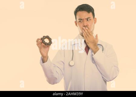Studio shot of young Asian man doctor holding donut while looking shocked Stock Photo