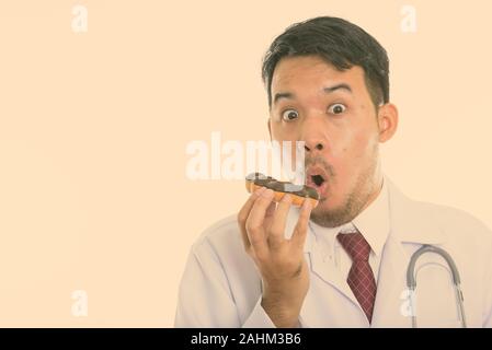 Studio shot of young Asian man doctor eating donut while looking shocked Stock Photo