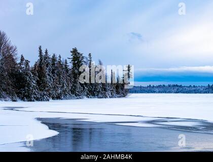 Snow-covered pine trees cover a small peninsula of woodland that extends out into a lake. Much of the lake is now frozen in winter, with liquid water Stock Photo