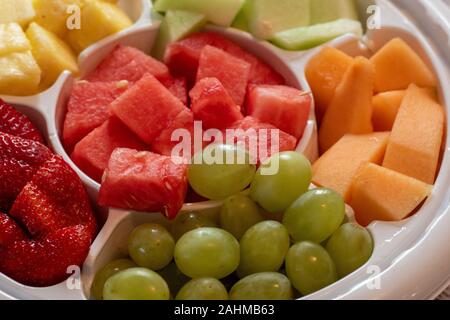A platter of fruit, consisting of watermelon, cantaloupe, green grapes, strawberries, pineapple and honeydew melon is served in a white plastic tray a Stock Photo