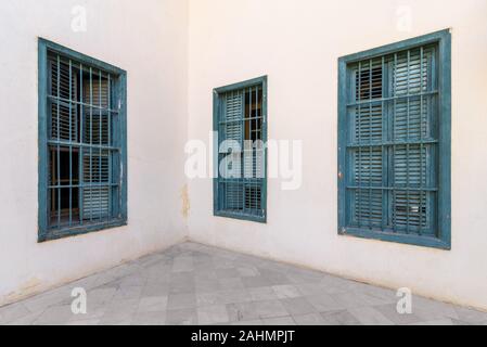 Corner white wall with three grunge windows with wooden green shutters and wrought iron bars and tiled white marble floor Stock Photo