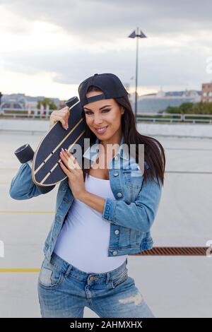 Happy young urban woman in denim holding longboard on shoulder Stock Photo