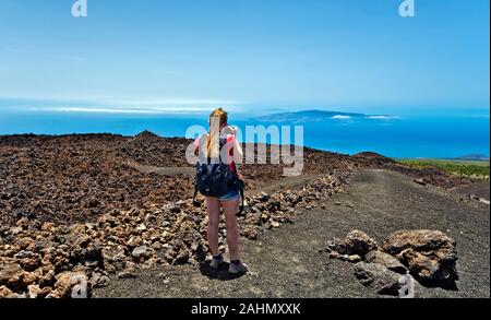 Young girl photographer, dressed for walking, takes photo of island landscape from Pico Viejo slope in Teide national Park in Tenerife Island, Spain. Stock Photo
