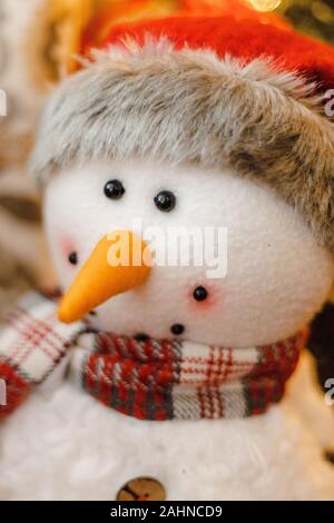 Portrait of a snowman stuffed animal plush smiling with Santa hat, red scarf, orange carrot nose with Christmas tree in the blurry bokeh background Stock Photo