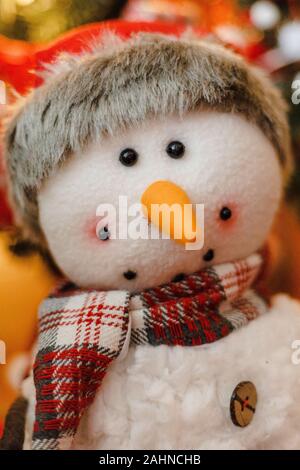 Portrait of a snowman stuffed animal plush smiling with Santa hat, red scarf, orange carrot nose with Christmas tree in the blurry bokeh background Stock Photo