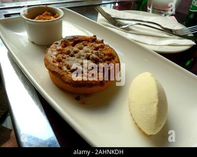 round shaped single serving Dutch apple pie. elongated white porcelain plate. dressing in small ceramic bowl on the side, white vanilla mousse cream. Stock Photo