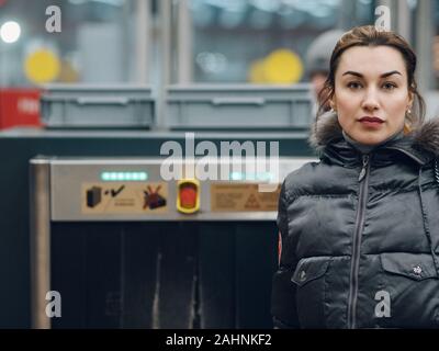 A young woman is looking at the camera. A brunette in demi-season clothes is standing near the security counter in the subway. In the background is a Stock Photo