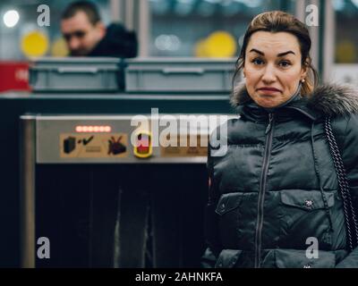 A young woman is looking at the camera. A brunette in demi-season clothes is standing near the security counter in the subway. In the background is a Stock Photo