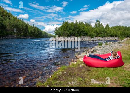 Young woman is relaxing in the red airlounger in the border of Fugga river in Hedmark county of Norway. Close to Akrestrommen village in Rendalen muni Stock Photo