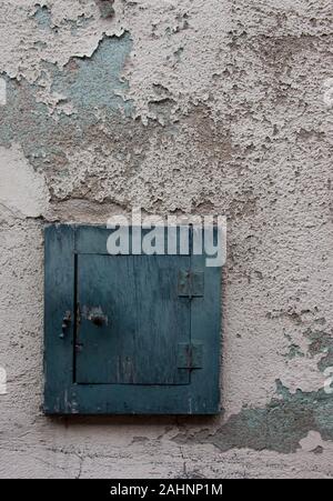 Small blue shuttered window on white peeling paint wall Stock Photo