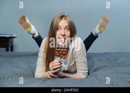 Funny emotional girl holding milk chocolate with whole nuts in her hand. Cheerful young woman lying on the bed and eating chocolate bar Stock Photo