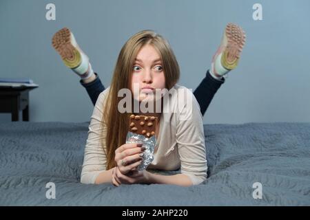 Funny emotional girl holding milk chocolate with whole nuts in her hand. Cheerful young woman lying on the bed and eating chocolate bar Stock Photo