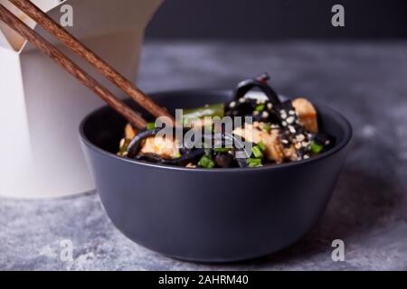 Black squid ink udon noodles with chicken meat on the black bowl with chopsticks on dark stone background and paper box nearby. Take out food concept Stock Photo