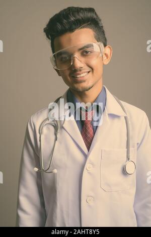 Studio shot of young Indian man doctor wearing protective glasses against gray background Stock Photo