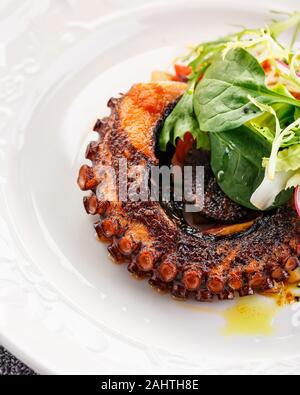 Octopus tentacle fried in a pan with new potatoes and calamata olives, garnished with green salad and spinach. Restaurant dish. Close-up Stock Photo