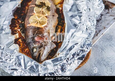 Baked rainbow raw trout with parsley and lemon in foil on a light baking sheet on a gray background. Healthy food. Cooking concept. Stock Photo