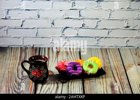 A black ceramic cup with fresh cappuccino and a plate with five donuts in colored glaze on a wooden table against a stone wall. Close-up. Stock Photo