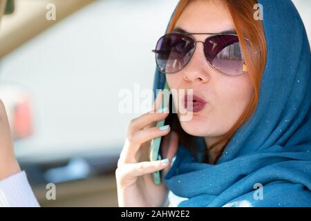 Fashionable woman driver in scarf and sunglasses talking on mobile phone while driving a car. Stock Photo