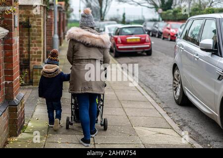 A mother pushing a pram, pushchair or stroller down a street while a young child walks alongside holding on Stock Photo