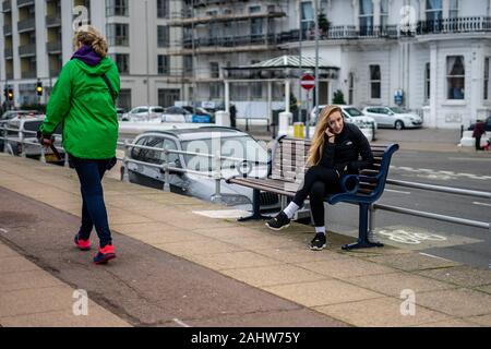 A woman using a mobile phone while sat alone on a bench as someone passes by Stock Photo