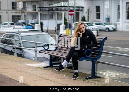 A teenage woman in sportswear sitting on a bench in the street talking on her mobile phone or cell phone Stock Photo