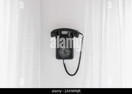 Vintage Black Rotary Phone hanging on Wall, flanked by White Curtains Stock Photo