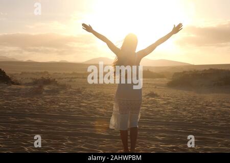 Sunset in the desert. Young woman with with raised arms wearing white dress walking in the desert dunes sand during sunset. Girl on golden sand on Cor