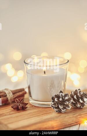 White burning candle with decorations and garland lights in bokeh on wooden table. Cozy home and hygge concept Stock Photo