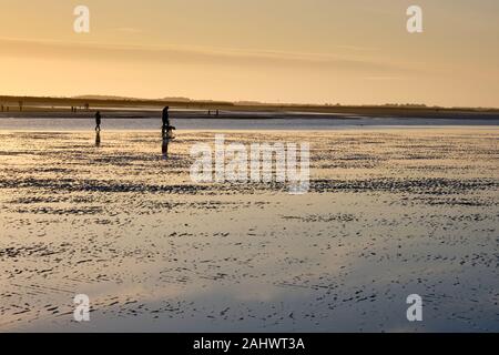 Figures of dog walkers silhouetted by the golden setting evening sun on a winter time Brancaster Beach on the North Norfolk Coast England Stock Photo