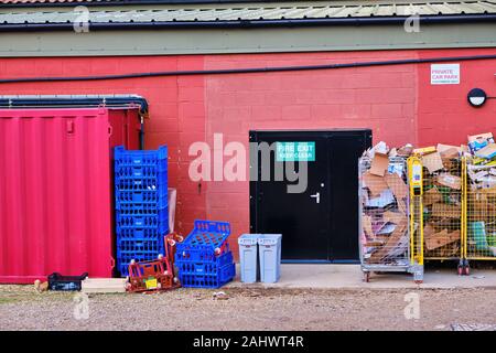 Black fire exit door in red painted building wall with sign keep clear surrounded by waste card and stacked boxes bins trash cans Stock Photo