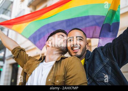 Portrait of young gay couple embracing and showing their love with rainbow flag at the street. LGBT and love concept. Stock Photo