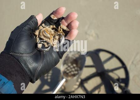 Metal detectorist holds jewelry found by metal detector on beach in Florida with detector coil and scoop in background on sand. Stock Photo