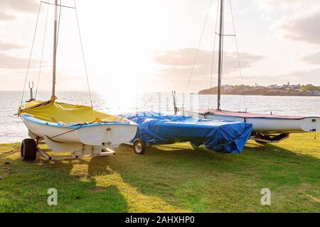 Sailing boats parked on grass next to the clubhouse, with early morning light shining across the ocean Stock Photo
