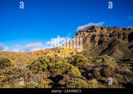 Spain, Tenerife, Fog clouds and moon moving over red rocky mountains covered by some green plants in caldera desert nature landscape with blue sky Stock Photo