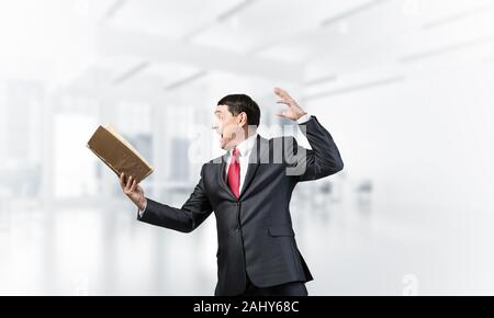 Expressive businessman holding open book. Portrait of adult man in business suit and tie standing in blurred office interior. Excited manager shouting Stock Photo