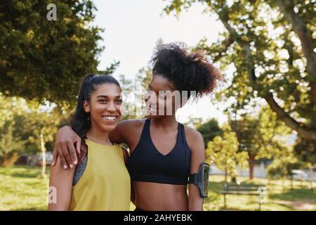 Smiling young women in sportswear standing arm in arm together at park - smiling multiethnic fitness friends in the park Stock Photo