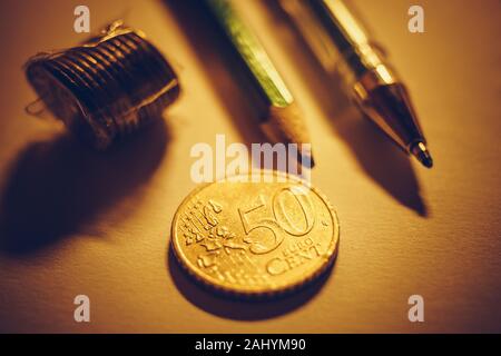 Golden 50 euro cent coin, stack of metal coins, pen and pencil on the paper table. Stock Photo