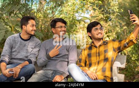 Group of friends taking a self portrait in the park - Concept of young people addicted to selfie - Three friends are sitting on bench in park and Stock Photo