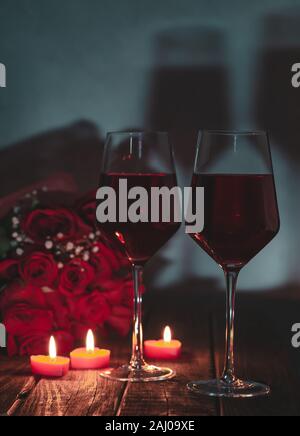 Dark scene of two glasses of wine with burning candles and bouquet of red roses in background on rustic wooden table Stock Photo