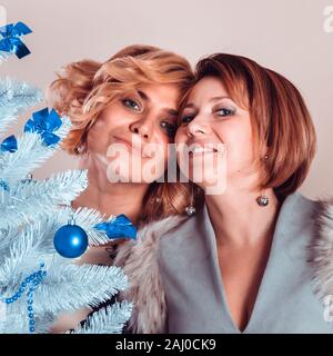 Elegant girlfriends in evening dresses and a fur collar stand nearby. Sisters hug. Young beautiful women smiling in anticipation of Christmas, gatheri Stock Photo