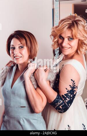 Elegant girlfriends in evening dresses and a fur collar stand nearby mirror. Sisters hug. Young beautiful women smiling in anticipation of Christmas, Stock Photo