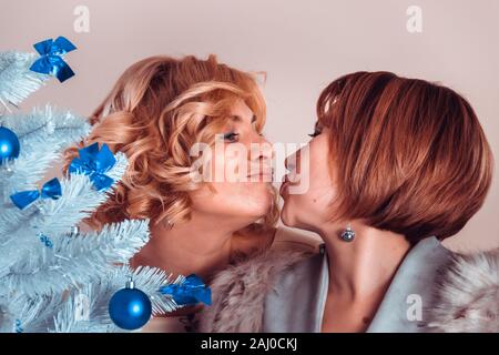 Elegant girlfriends in evening dresses and a fur collar stand nearby. Sisters hug. Young beautiful women smiling in anticipation of Christmas, gatheri Stock Photo